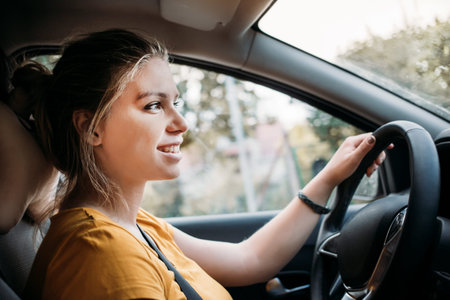 Young beautiful girl sitting behind the wheel of a car wearing a seat belt and looking at the road,...