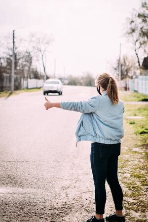 A young woman in a black medical mask with blond hair in a turquoise jacket stands with her back to the camera with her hand raised and catches the car on the road in the village. Quarantine concept.の写真素材