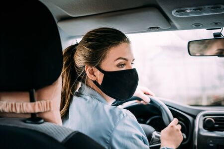 A young taxi driver woman in a black medical mask with blond hair and a turquoise jacket is driving a left-hand drive car. A woman looking at road. Masks protective. Quarantine. Virus. Pandemic.の写真素材
