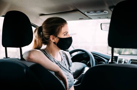 A young woman in a black medical mask with blond hair in a gray T-shirt is sitting in a left-hand drive car, put her hand on the seat. Portrait photo. Quarantine concept. Virus. Pandemic. Coronavirusの写真素材