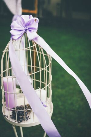 Decorative, white bird cage with a lilac bow on the top. Boquet of lavander and two candels inside. Close up. Wedding concept.の写真素材