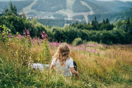 A girl in a white T-shirt and blond hair sits on a slope and enjoys the view of blooming mountains.の写真素材