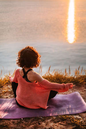 Vertical photo of a woman doing yoga at dawn and looking at the water and the suns reflection in the water. Pacification, balance, tranquilityの写真素材