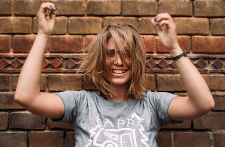 Happy cheerful female with short blond hair laughing and waving her hands near a brick wallの写真素材