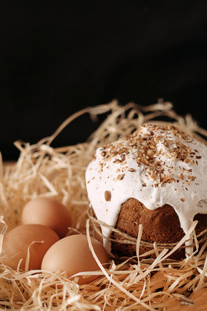Easter cake decorated with white icing next to chicken eggs in a straw on a black background.