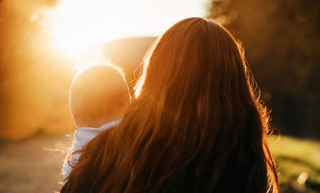 Blurred mom close up with child at sunset. Orange backlighting and bokeh. Mother and baby concept, inspiration. Back viewの写真素材