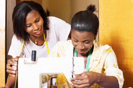The professional seamstress sews and his trainee assists from behind.