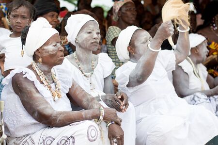 adzop?, ivory coast-31August 2016 Mature woman dressed in white with powder on the body, arm bracelet with ponytail in hand sitting both with arms closed at her side and in the background other women seated.のeditorial素材
