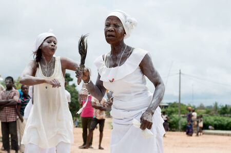 adzop?, ivory coast- August 31, 2016: Mature woman dressed in white necklace with ponytail neck in hand dancing singing accomgnated by a young woman in white dress, in the background of the young people standing.のeditorial素材