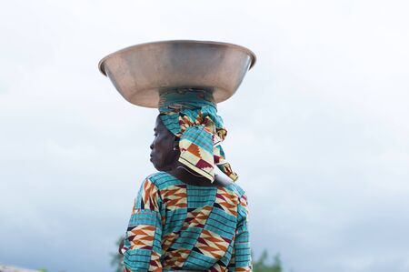 Adzope, Ivory Coast - June 10, 2017: Portrait of a woman in back in a camisole farcloth and head tied to the farcloth, loads a basin in alluminumのeditorial素材