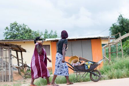 Adzop?, ivory coast - June 10, 2017: two young African women, one in black T-shirt and farcloth pushes a full wheelbarrow and the other in a farcloth dressのeditorial素材