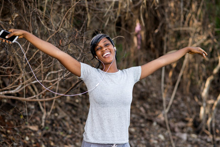 young girl in sportswear standing near a bush with arms outstretched and with earphones, listening...