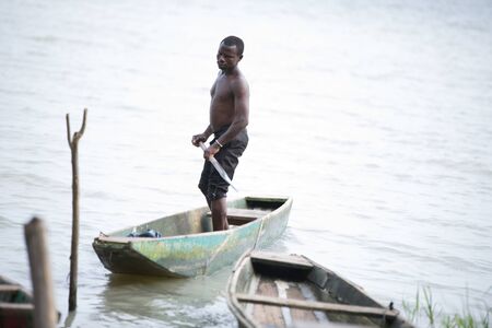 Abidjan, Ivory Coast -11 February 2018: Shirtless young man standing in a dugout canoe on the water slowly moving towards the shore.のeditorial素材