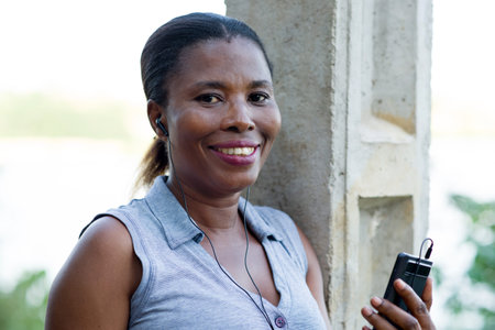 young woman smiling with a mobile phone and leaning against an electric pole outside ..