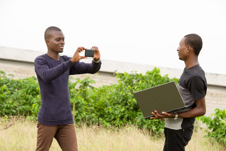 Young people standing in grass one with laptop in hand while the other makes him photo with mobile...