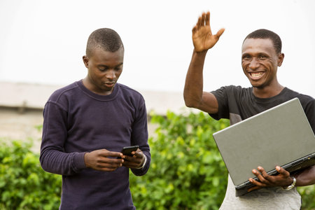Young people standing outdoors one with mobile phone, the other with computer and laughing while...