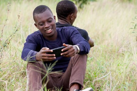 Close up of two young men leaning against each other and sitting on the grass in the park. one seen from behind and the other seen from the front uses a mobile phone. Concept of friendship men.の写真素材