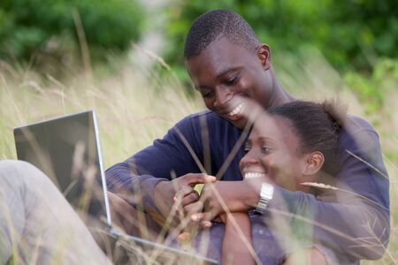 concept of love and people - happy teenage couple lying on the grass in front of a laptop, laughing and sharing moments of happiness while looking on smart phone in the summer.の写真素材