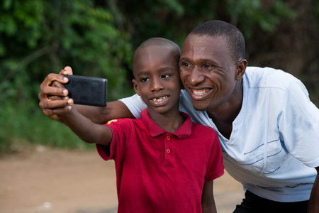 little boy in red t-shirt standing near his father and looking at mobile phone laughing.