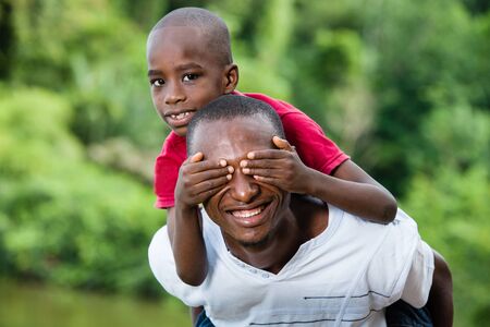 child sitting on his father's neck is holding his eyes closed.Happy young father and son having fun and enjoying the fresh air in nature.の写真素材