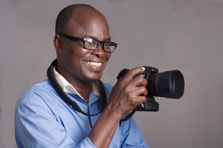 African man standing in blue shirt on gray background looking ahead with camera in hand.