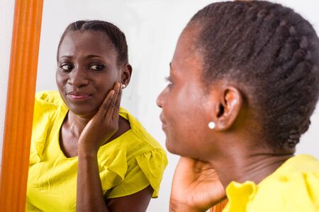 African girl standing in yellow tank looking at herself in a mirror smiling.の写真素材