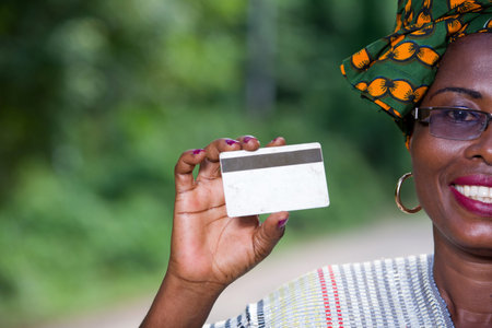 portrait of happy african woman in traditional clothes standing in nature showing a credit card,