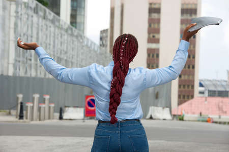 Happy businesswoman standing back with long red braids, holding a document with outstretched arms and relaxing outside offices feeling ecstatic on the street in summer. Copy spaceの写真素材