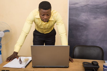 Charming businessman leaning on a conference table in the office and looking on laptop
