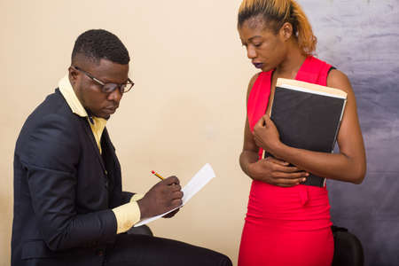 young businessman sitting in office suit writing and his secretary standing with documents.の写真素材
