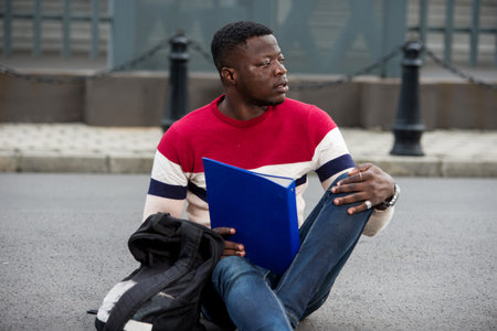 Handsome college man reading notes in notebook on campus.Happy student learning outdoors sitting...