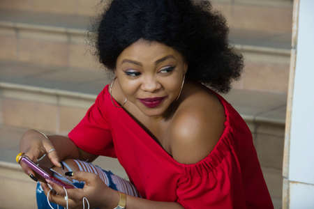 Funny african woman sitting on stairs listening to music with headphones and a smart phone outdoors.の写真素材
