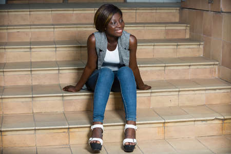 smiling young woman with elegant jacket and blue jeans sitting on the stairs.の写真素材