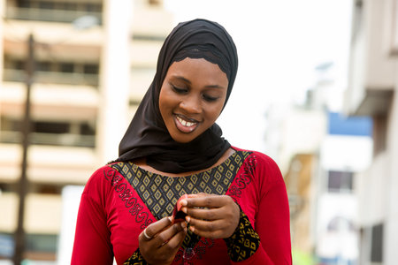 young muslim woman standing outdoors looking at lipstick smiling.
