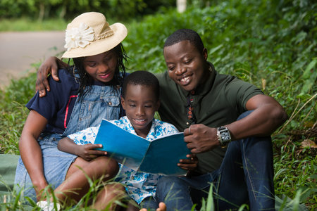 a couple sitting in the grass watching their child read with a smile.