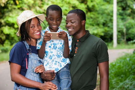 a happy family standing in a park watching mobile phone with their son smiling.の写真素材