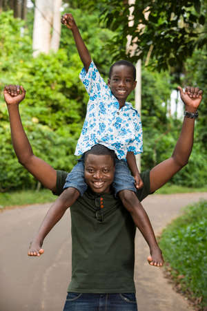 a young man standing in a park with his son on his neck doing gestures while smiling.の写真素材