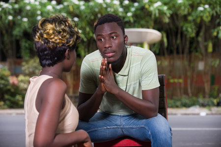 young man in t-shirt sitting outdoors looking at his girlfriend with clasped hands.の写真素材