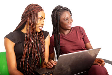 two beautiful young women sitting on a chair isolated on white background with laptop and looking together at something smiling.の写真素材