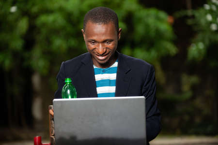 young man sitting on a bench in a park and drinking juice in a green plastic bottle using a laptop computer smilingの写真素材