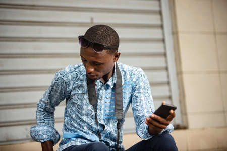 handsome young african photographer sitting outside holding a mobile phone and looking at camera smilingの写真素材