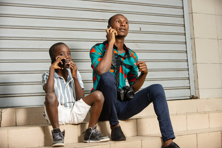 Handsome young man and his pre-teen son sitting on the steps of a stairs next to each other using a...