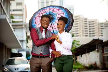 portrait of a young african couple standing under an umbrella outdoors in the street while smilingの写真素材