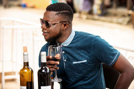 handsome young man wearing sunglasses and sitting aside alone outdoors and drinking a glass of red wineの写真素材
