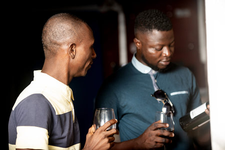 two handsome young african men standing holding a glass of red wine and looking at the bottle of...