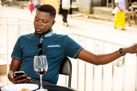 handsome young man sitting outdoors using a mobile phone while having a glass of red wine on the...