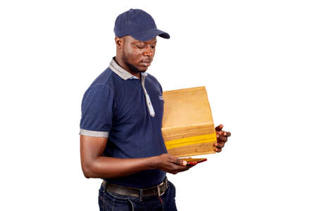 young delivery man standing in blue cap on white background carrying a package looking for contact in his mobile phone.の写真素材