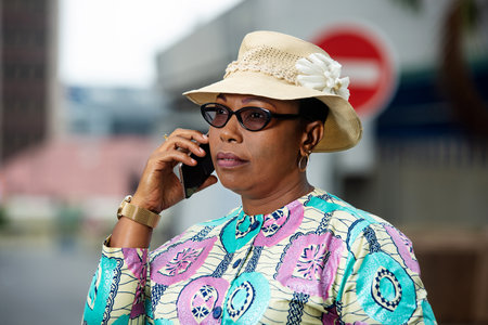 beautiful adult businesswoman wearing glasses and a Belgian hat walking and talking using a mobile phone in the streetの写真素材