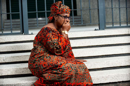 beautiful mature woman wearing glasses sitting on stairs outside is shocked and putting her hand on her mouthの写真素材