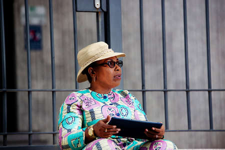 beautiful african adult woman sitting holding her tablet with a on her head wearing a beautiful loincloth and glasses pharmaceutical and with the handle a beautiful watchの写真素材